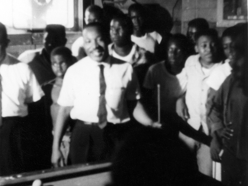 The Rev. Martin Luther King Jr. is surrounded by admirers in a Neshoba County pool hall, where he spoke about registering to vote and supporting the Mississippi Freedom Democratic Party, July 1964. A sign in the upper left corner of the photo says, "No Bad Language." (Photo used with the permission of the Rev. Ed King, University Press of Mississippi and the Mississippi Department of Archives and History)